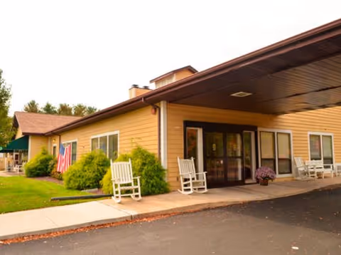 Exterior view of a single-story building with a covered entrance, beige siding, several windows, and white rocking chairs on the porch. There are green bushes and an American flag near the entrance.