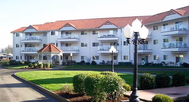 Front exterior of a three-story senior living building with balconies, a gazebo-style entrance, lamp posts, and landscaped lawn.