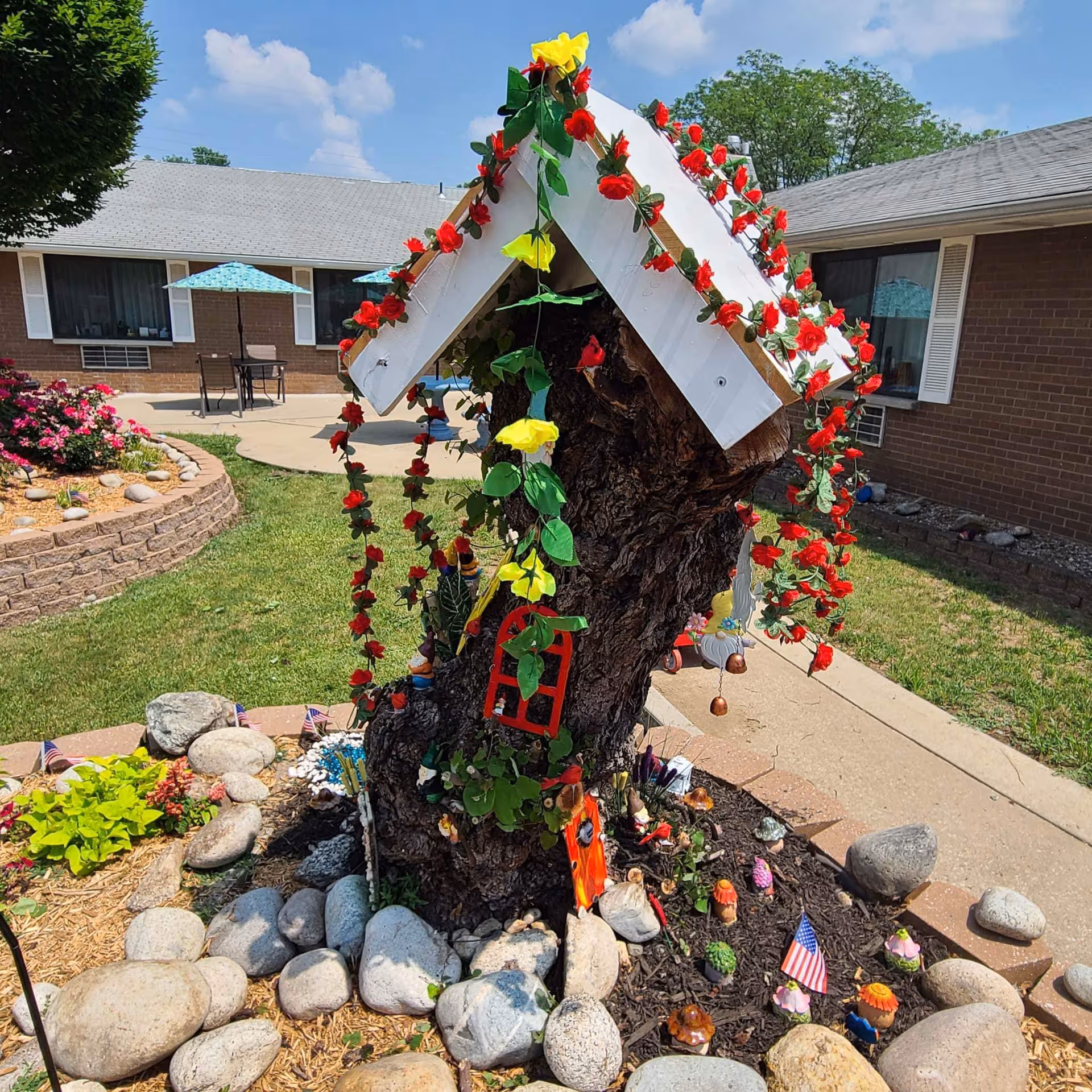 A small decorative garden area featuring a tree stump with a white roof adorned with red and yellow artificial flowers. The stump is decorated with miniature items including a red window frame, small figurines, and tiny American flags. Surrounding the stump are rocks and mulch, with a brick building and patio furniture with blue umbrellas visible in the background under a partly cloudy sky.