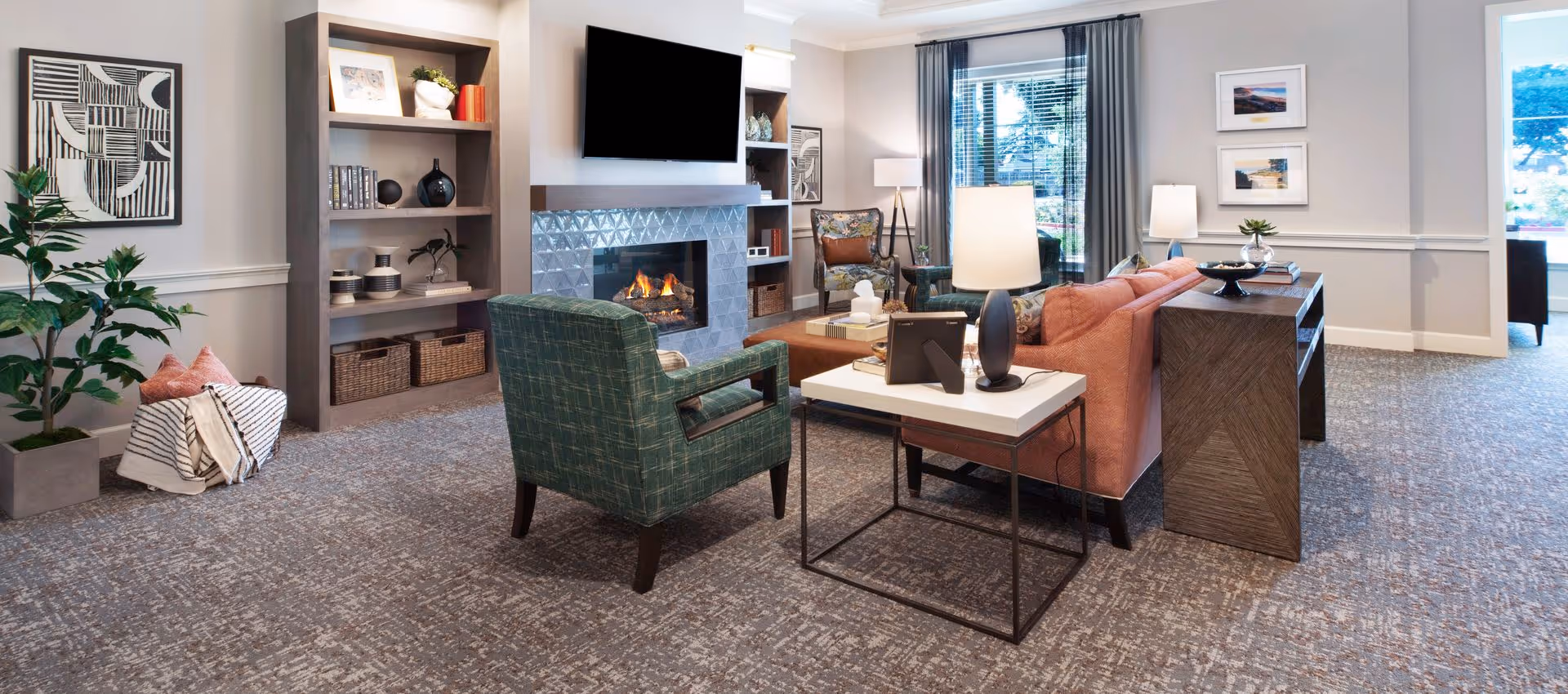 A cozy living room area with a green armchair, a pink sofa, a white side table with a lamp, and a wooden console table. There is a fireplace with a TV mounted above it, built-in shelves with decorative items, a large window with gray curtains, and framed artwork on the walls. The room has carpeted flooring and a potted plant in the corner.