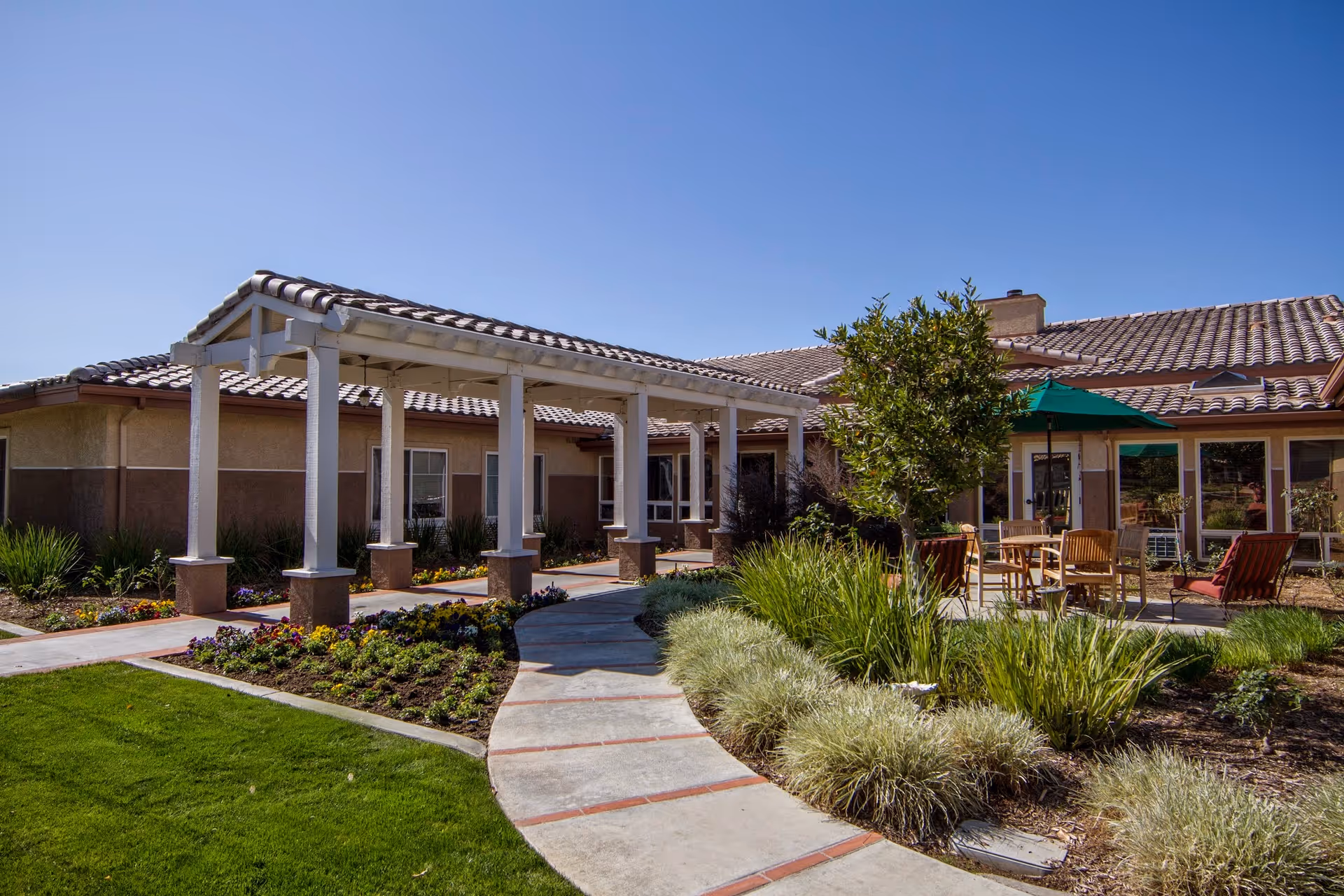Outdoor view of a senior living facility with a covered walkway supported by white columns, landscaped garden beds with flowers and shrubs, a curved concrete pathway, and a patio area with wooden chairs and tables under a green umbrella. The building has a tiled roof and beige walls under a clear blue sky.