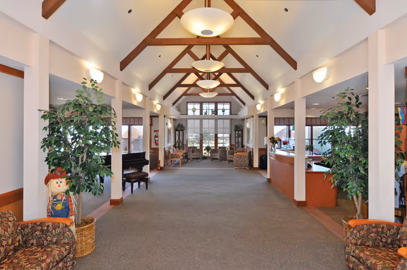 Spacious interior common area with high vaulted ceiling featuring wooden beams and hanging light fixtures. The room is carpeted and furnished with patterned armchairs, potted plants, and a piano on the left side. Large windows at the far end allow natural light to fill the space. A reception desk is visible on the right side.