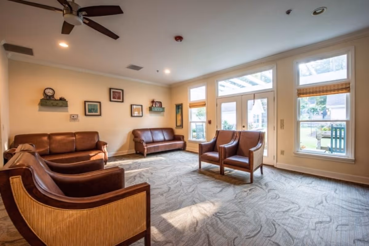 Bright common living room with leather sofas and armchairs, carpeted floor, ceiling fan, and large windows and doors looking out to a courtyard.