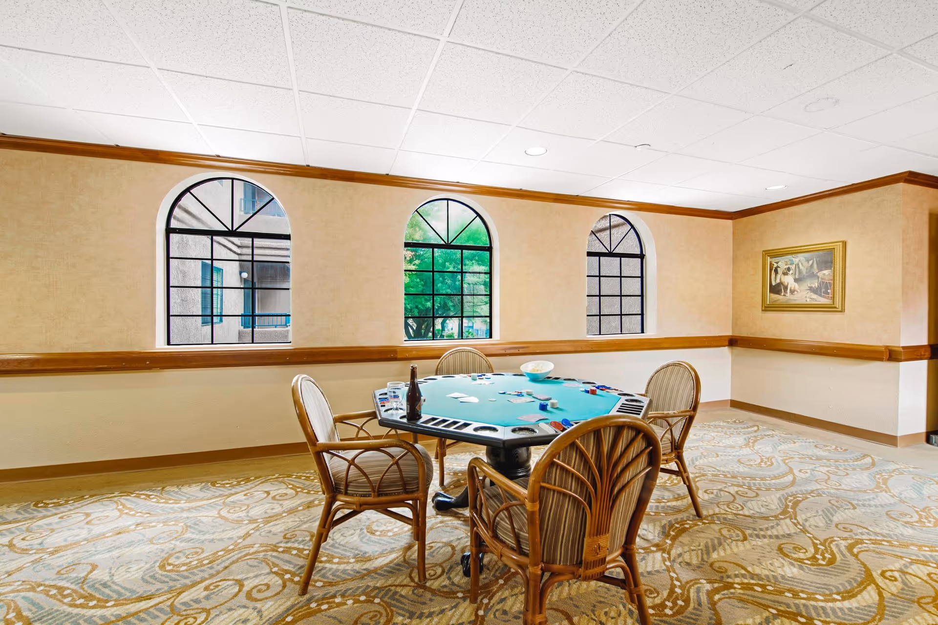 A well-lit room with three arched windows showing an outdoor view. In the center, there is a green felt poker table with playing cards, poker chips, and a bottle on it. Four wicker chairs with cushions surround the table. The room has beige walls with wooden trim and a patterned carpet. A framed painting hangs on the right wall.