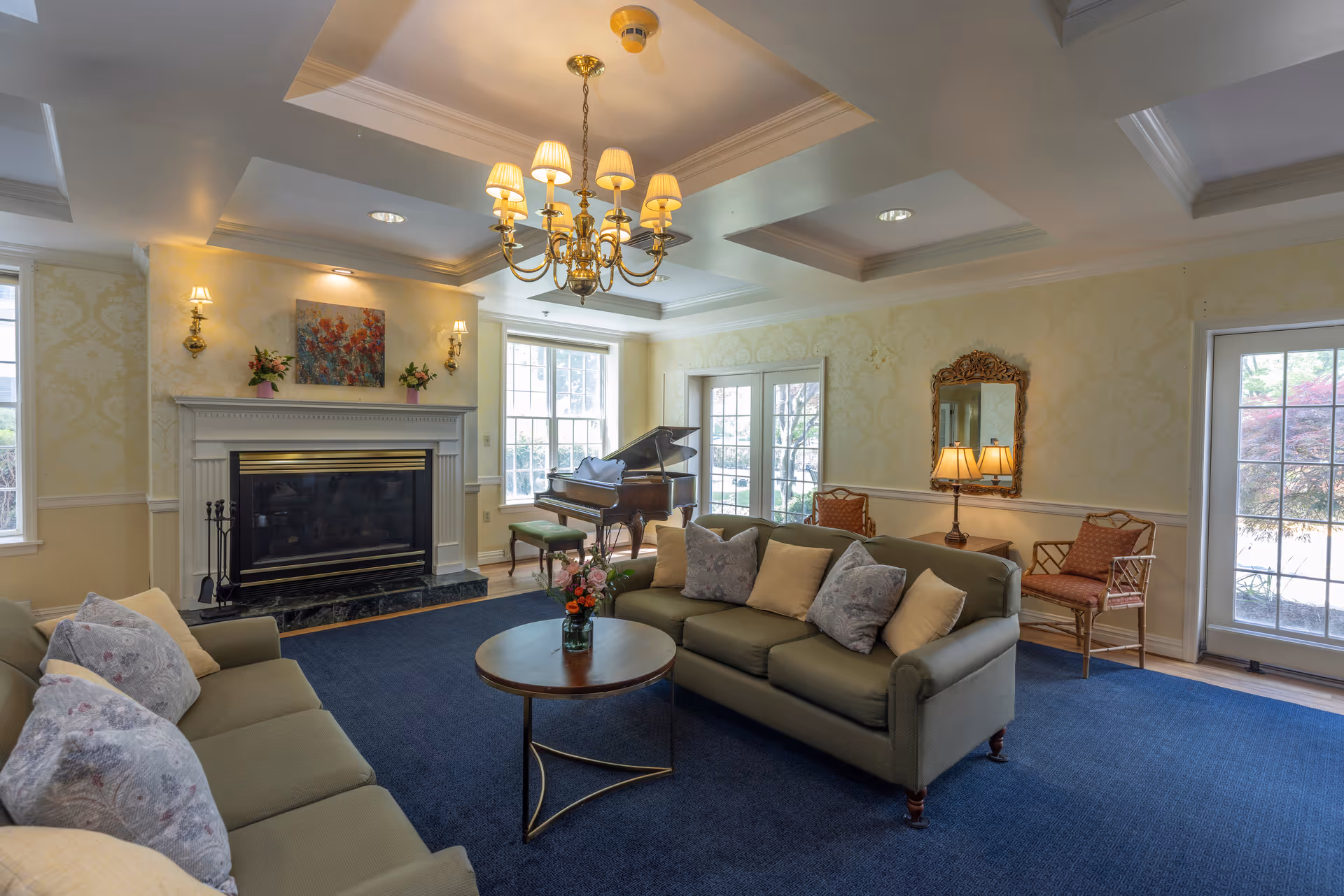 A cozy living room with two green sofas adorned with patterned and plain cushions, a round wooden coffee table with a flower vase, a grand piano near large windows, a fireplace with floral artwork and wall sconces above it, a chandelier hanging from a coffered ceiling, and a side table with a lamp and two chairs next to a glass door leading outside.
