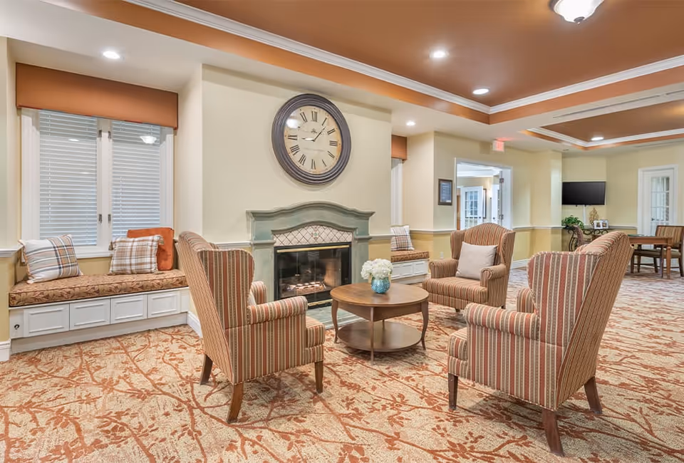 A cozy senior living facility common area with four striped armchairs arranged around a round wooden coffee table with a flower vase. There is a fireplace with a large clock above it, built-in window seats with cushions, and warm lighting from recessed ceiling lights. The carpet has a floral pattern and there is a TV mounted on the far wall.