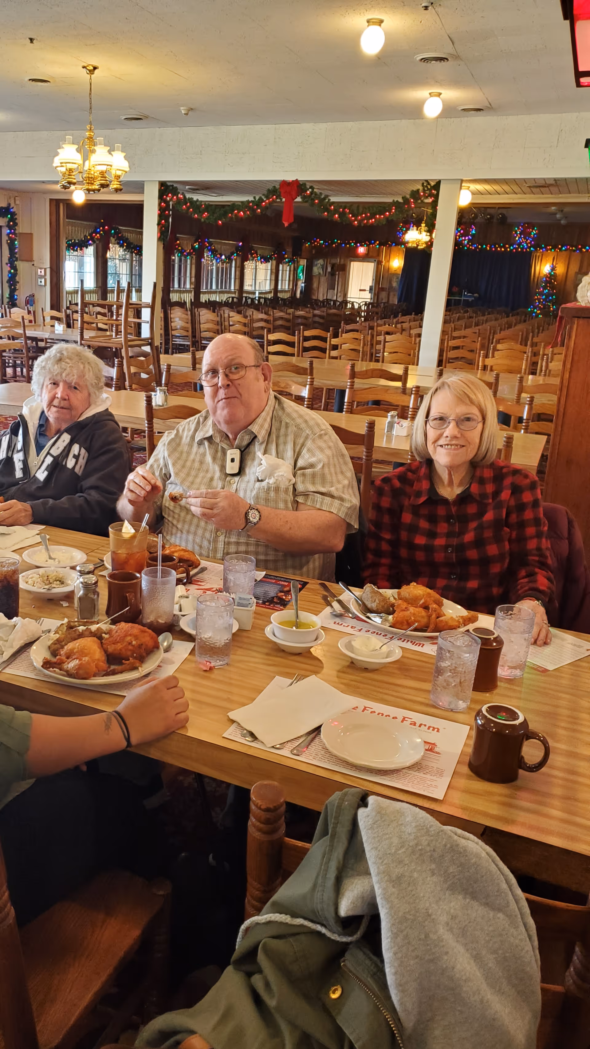 Three older adults sit at a long, decorated dining table in a communal dining room, enjoying a meal.