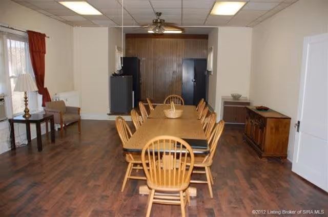 A dining room with a long wooden table surrounded by wooden chairs. The room has hardwood flooring, a ceiling fan, and a lamp on a side table near a window with red curtains. There are cabinets and appliances against the far wall.