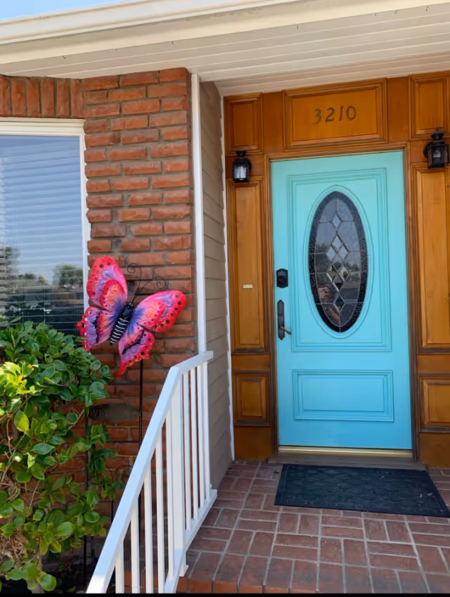 Front entrance of a house with a bright turquoise door featuring an oval glass panel with decorative patterns. The door is framed by wooden panels and has the number 3210 above it. There are two black wall-mounted lantern lights on either side of the door. To the left, there is a brick wall with a window and a green shrub. A large decorative pink and purple butterfly is mounted on a metal stand near the shrub. The entrance has a white railing and a black doormat on the brick floor.