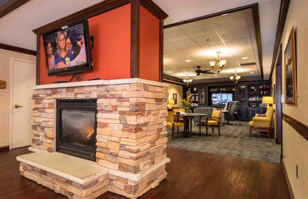 Cozy senior living common room with a stone fireplace and wall-mounted TV opening into a seating area with tables, chairs, and bookshelves.
