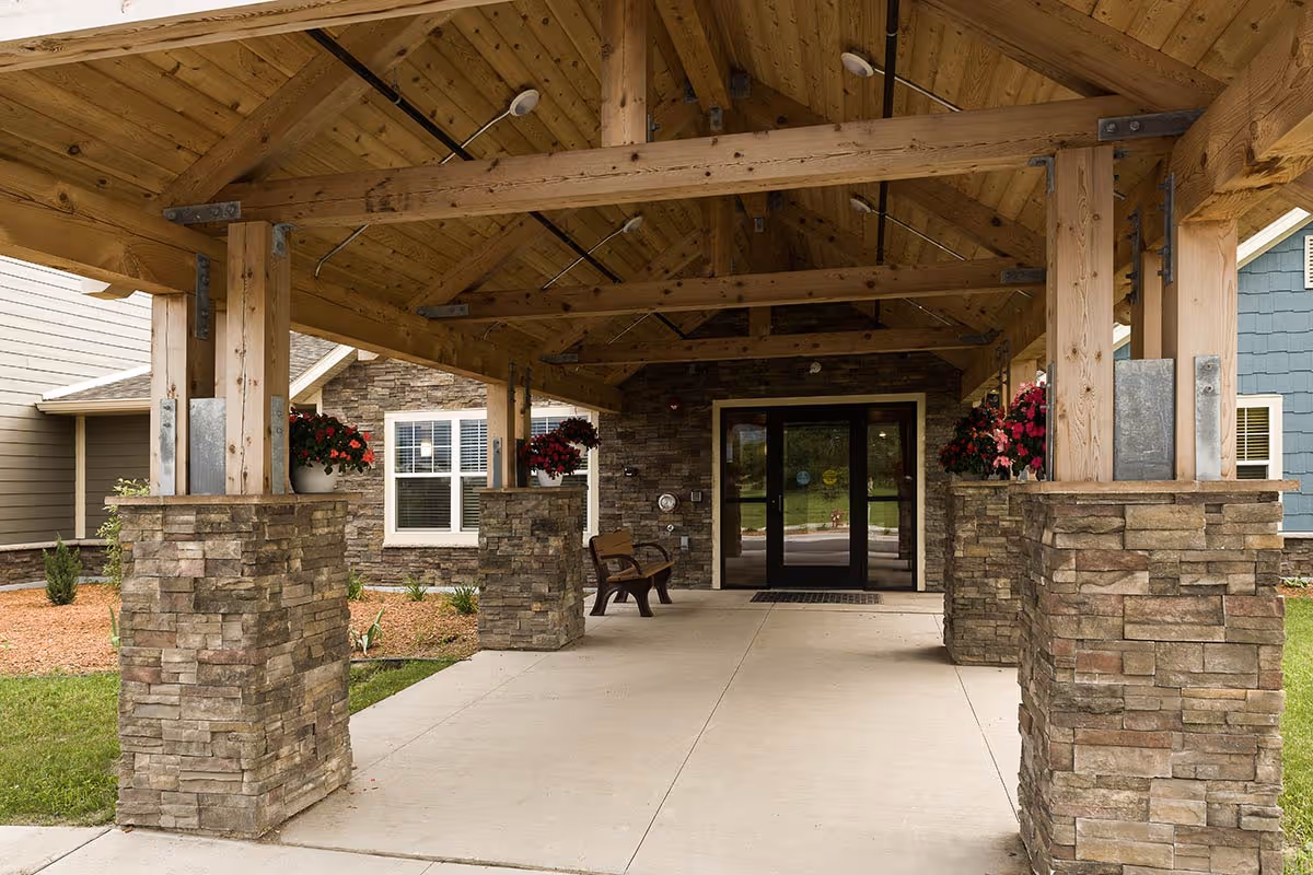 Covered entrance to a senior living facility with wooden beams and stone pillars, flower pots with red flowers on the pillars, a bench near the glass double doors, and a well-maintained pathway leading to the entrance.