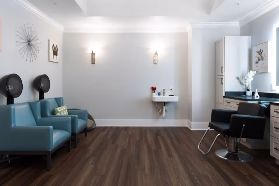 A modern salon room with two blue salon chairs with hair dryers on the left, a black salon chair on the right in front of a counter with cabinets, a small wall-mounted sink in the center, and decorative wall art and lighting.