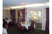A dining room with round tables covered with white tablecloths and surrounded by purple cushioned chairs. The room has beige walls with white trim, purple curtains on the windows, and two chandeliers hanging from the ceiling. There is a doorway leading to another room with additional seating and windows letting in natural light.