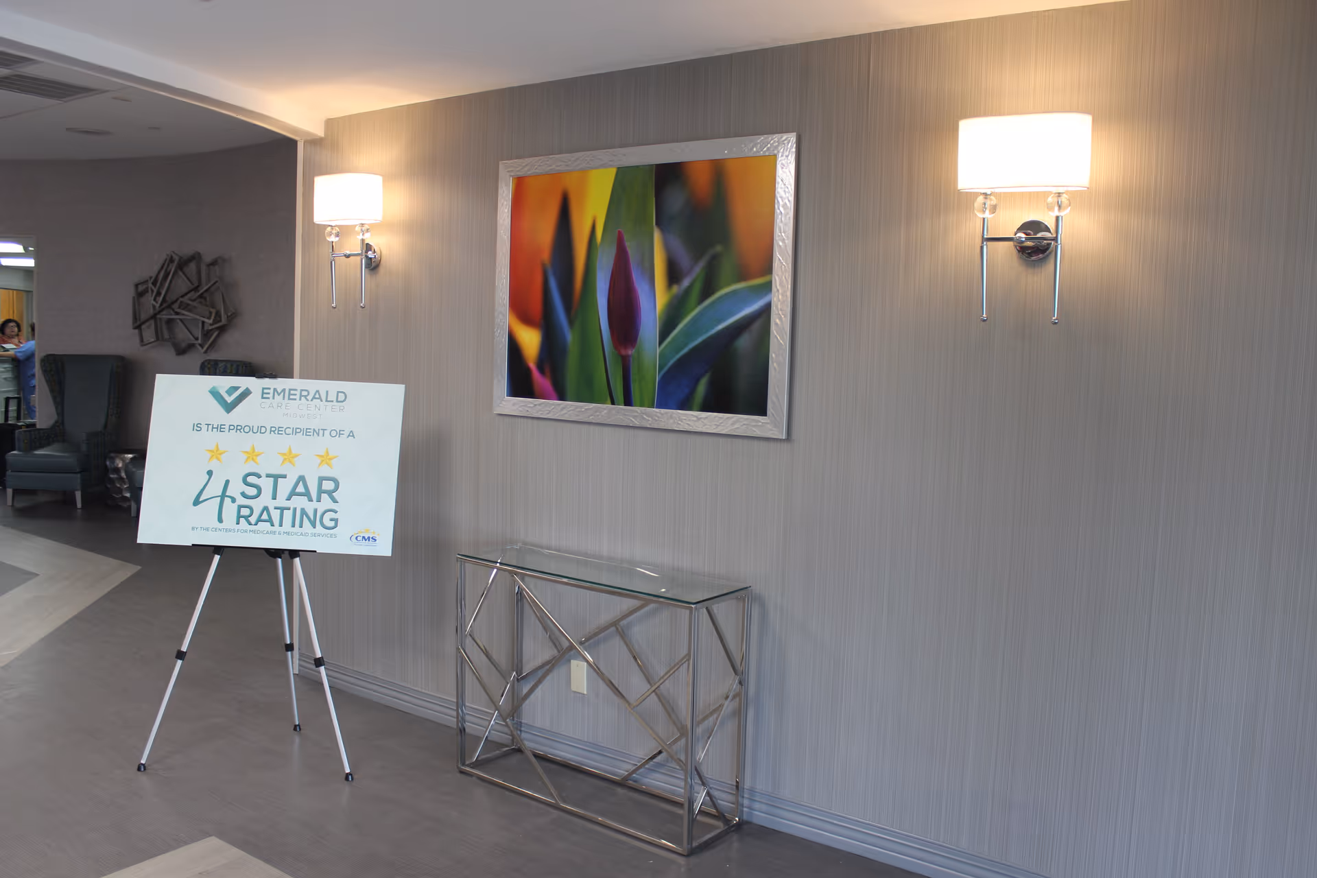Lobby interior with a framed floral artwork, two wall sconces, a glass console table, and a sign on an easel announcing Emerald Care Center's 4-star rating.