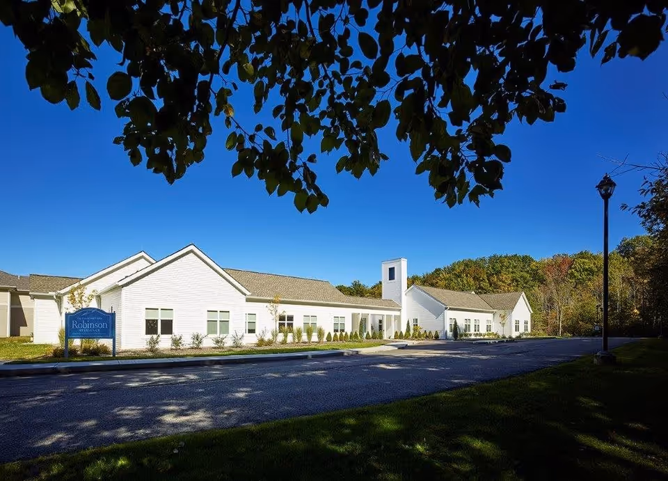 Exterior view of a single-story white building with a pitched roof under a clear blue sky. The building is surrounded by a paved driveway, green grass, and trees. A blue sign near the building reads 'Robinson Residence'. Tree branches with green leaves frame the top of the image.