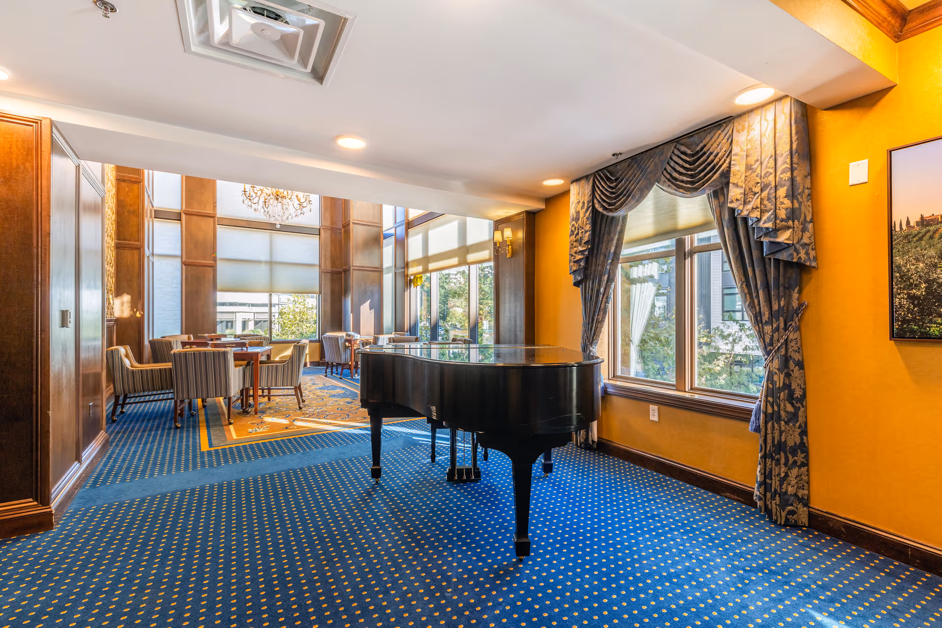 Interior room with a grand piano on a blue carpet with yellow dots, large windows with patterned blue curtains, and a seating area with tables and chairs in the background. The room has yellow walls and wood paneling, with a chandelier hanging in the seating area.