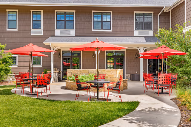 Outdoor patio area at Balfour Littleton with several round tables and red chairs, each shaded by a red umbrella. The patio is adjacent to a two-story building with brown siding and multiple windows. Green grass and landscaping surround the paved patio area.