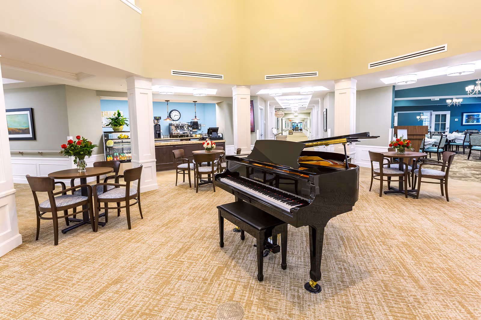 Interior of a senior living facility common area with a black grand piano in the center, surrounded by small round tables with chairs and flower vases. In the background, there is a coffee station with a clock on the wall and a hallway leading further into the facility.