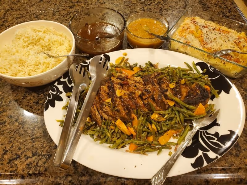 A meal spread on a granite countertop featuring a large white and black patterned plate with sliced roasted meat and green beans with carrots, a bowl of white rice, two glass bowls containing brown and yellow sauces, and a rectangular glass dish with a baked casserole topped with cheese.