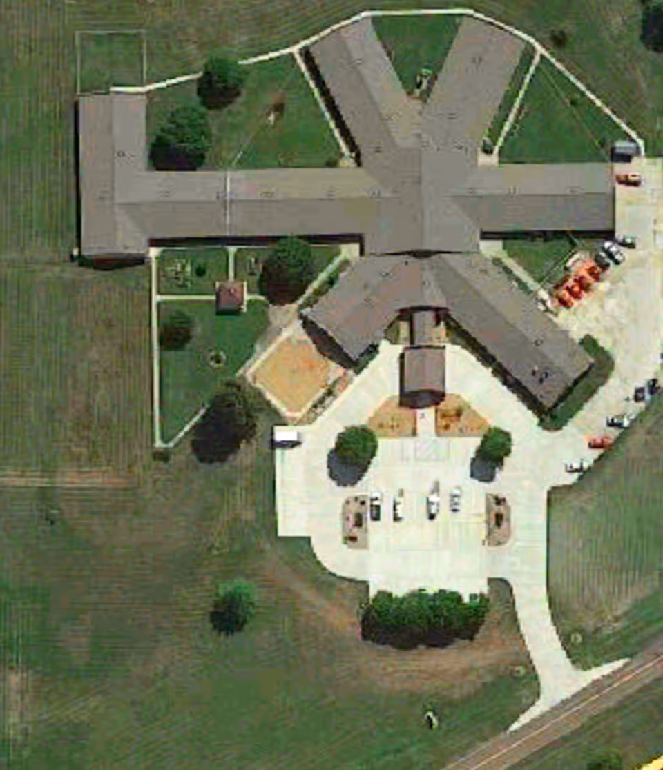Aerial view of the General Baptist Nursing Home of Linn, MO showing the building layout, surrounding green lawns, parking area with several vehicles, and pathways leading to the entrance.