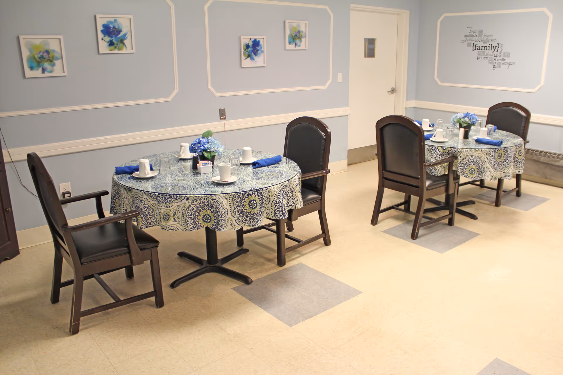 Two round tables with patterned tablecloths, place settings and floral centerpieces in a small dining room with blue walls and framed artwork.