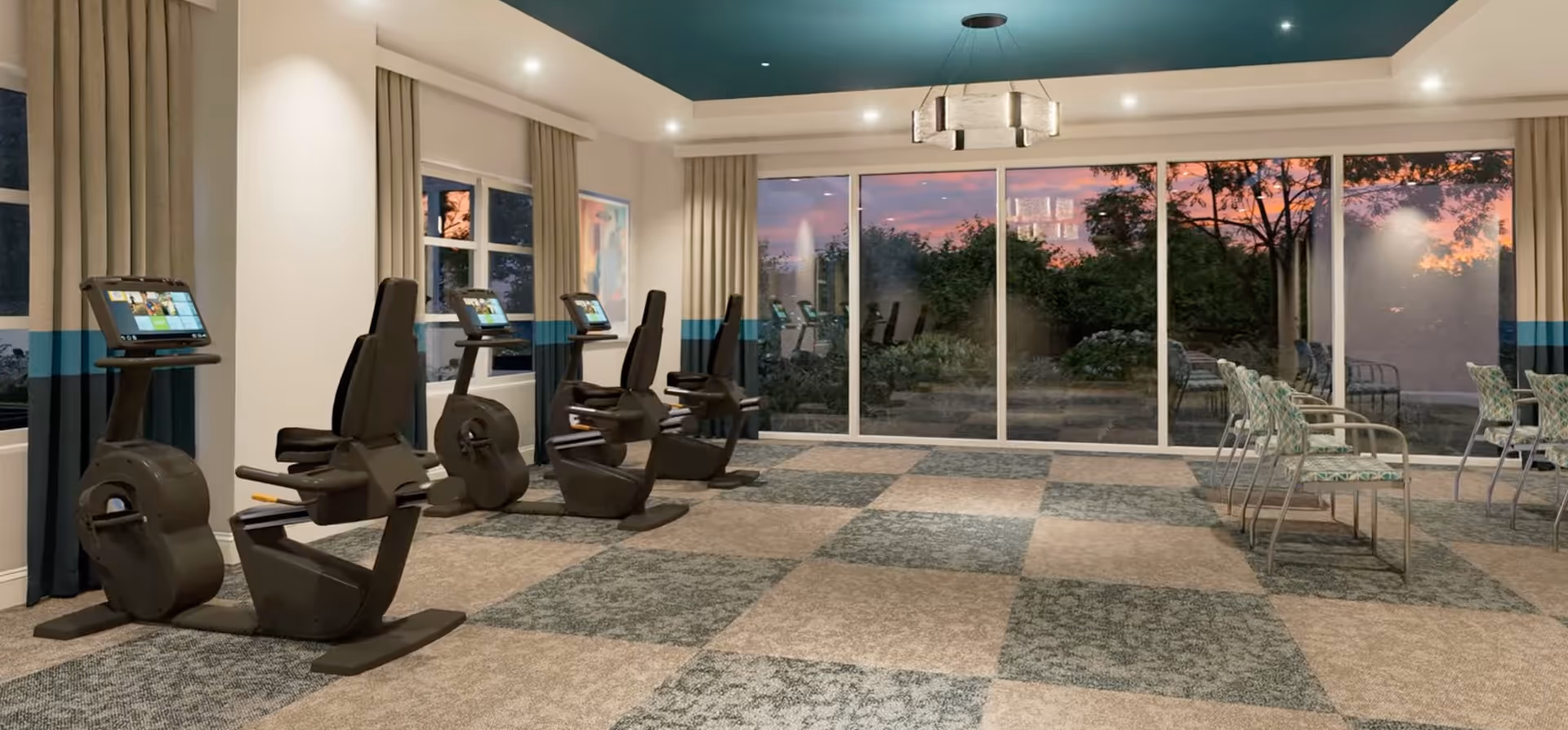 Fitness room with recumbent exercise bikes and chairs facing large floor-to-ceiling windows overlooking a sunset garden.
