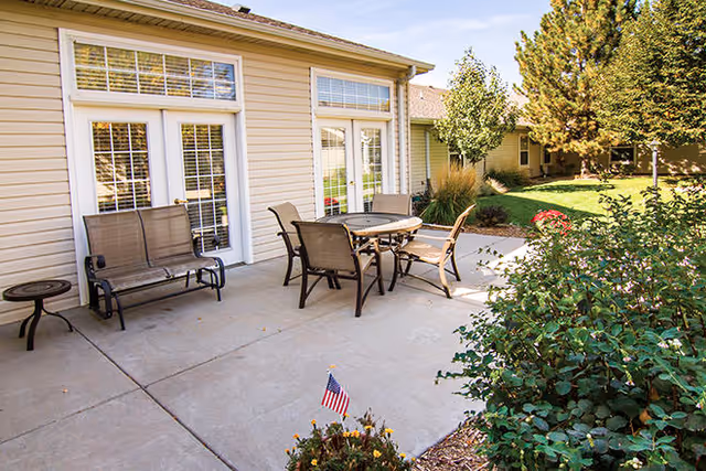 Outdoor patio area at Brookdale Brighton with a round table and four chairs, a bench, and a small side table on a concrete surface. The patio is adjacent to a building with large windows and glass doors. Surrounding the patio are green bushes, trees, and a well-maintained lawn under a clear sky.