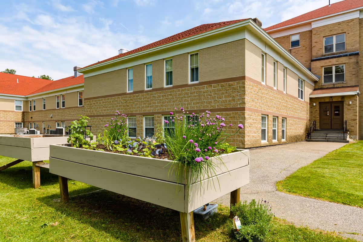 Exterior view of a brick senior living facility with raised wooden garden beds, a grassy lawn and a walkway leading to the main entrance.