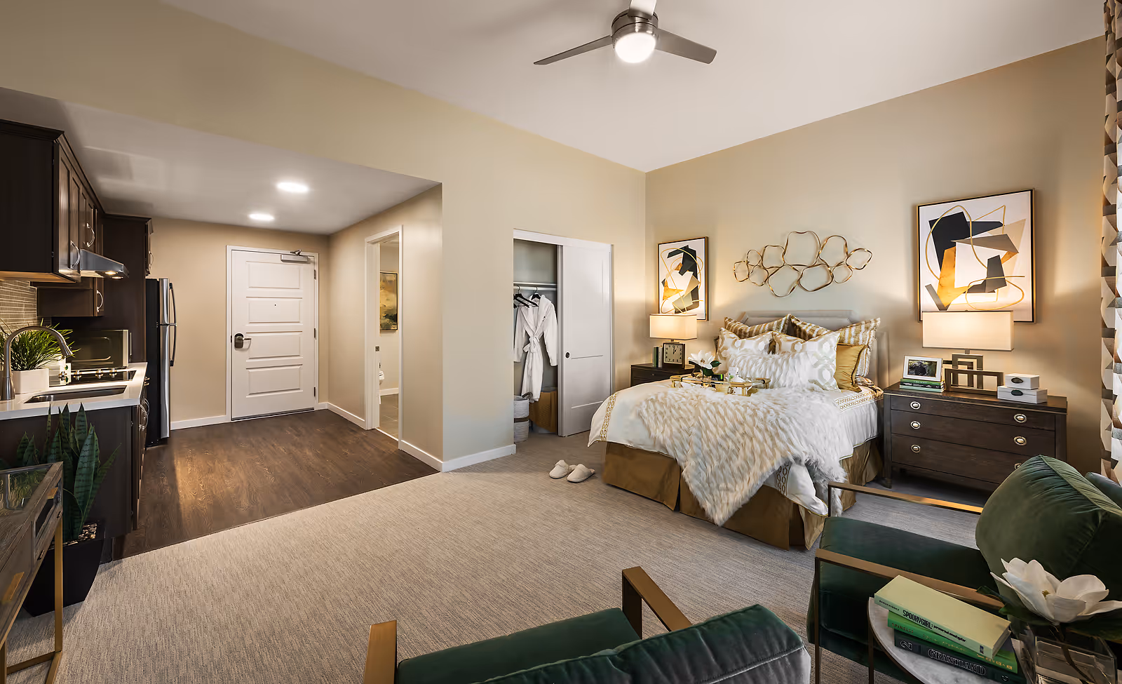 A modern studio apartment interior featuring a cozy bed with multiple pillows and a soft throw blanket, two bedside tables with lamps and abstract artwork above, a small open closet with hanging robes, a kitchenette with dark cabinets and stainless steel appliances, and a seating area with green velvet chairs and a small table with books and a flower. The room has beige walls, carpeted flooring in the bedroom area, and wood flooring near the entrance and kitchenette.