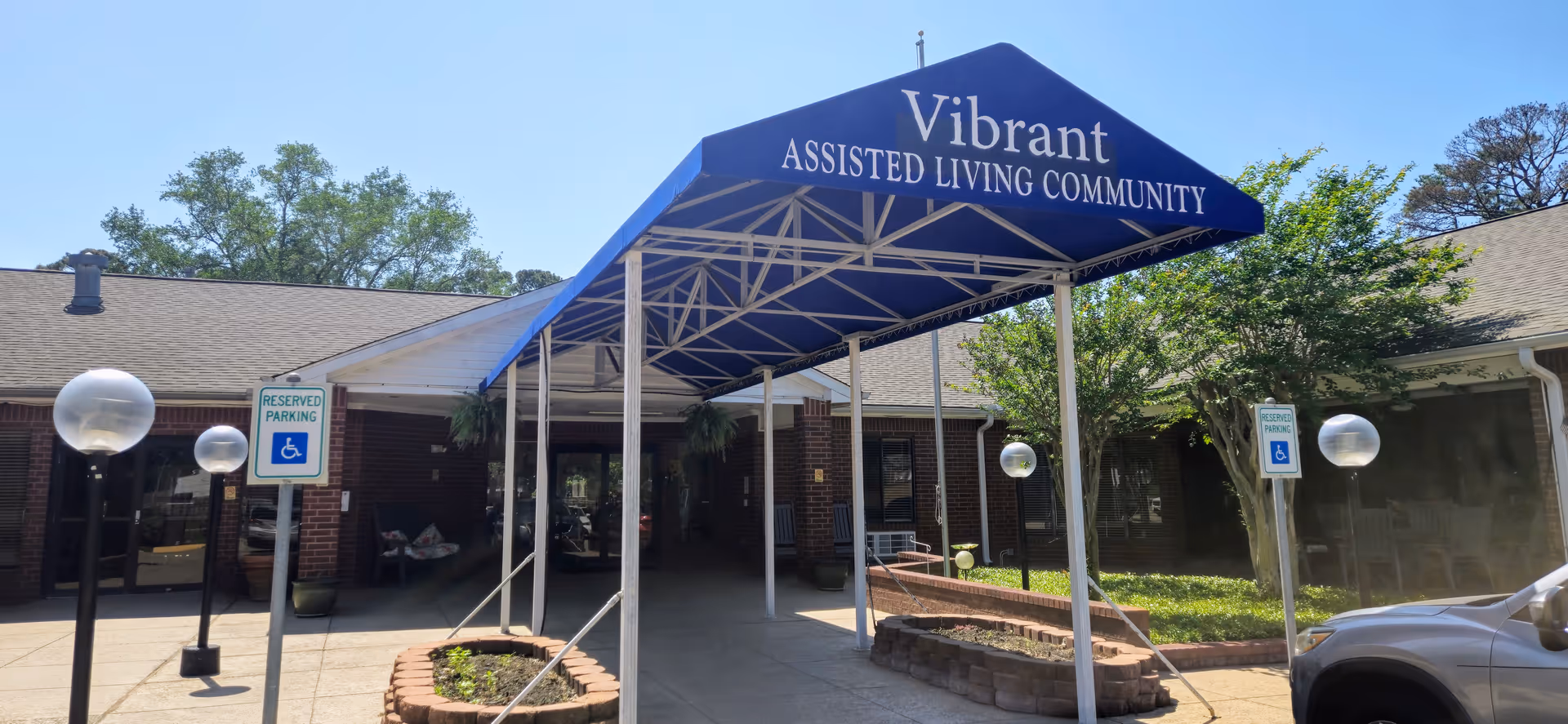 Entrance to Vibrant Assisted Living Community with a blue canopy displaying the facility name. The building is single-story with brick walls and a shingled roof. There are reserved parking signs for handicapped parking and globe-shaped outdoor lights along the walkway leading to the entrance.