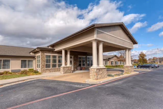 Exterior view of Cedarhurst Senior Living of Springfield, MO showing the building entrance with a covered drop-off area supported by columns, stone and beige siding, windows, and a parking lot with cars under a partly cloudy sky.