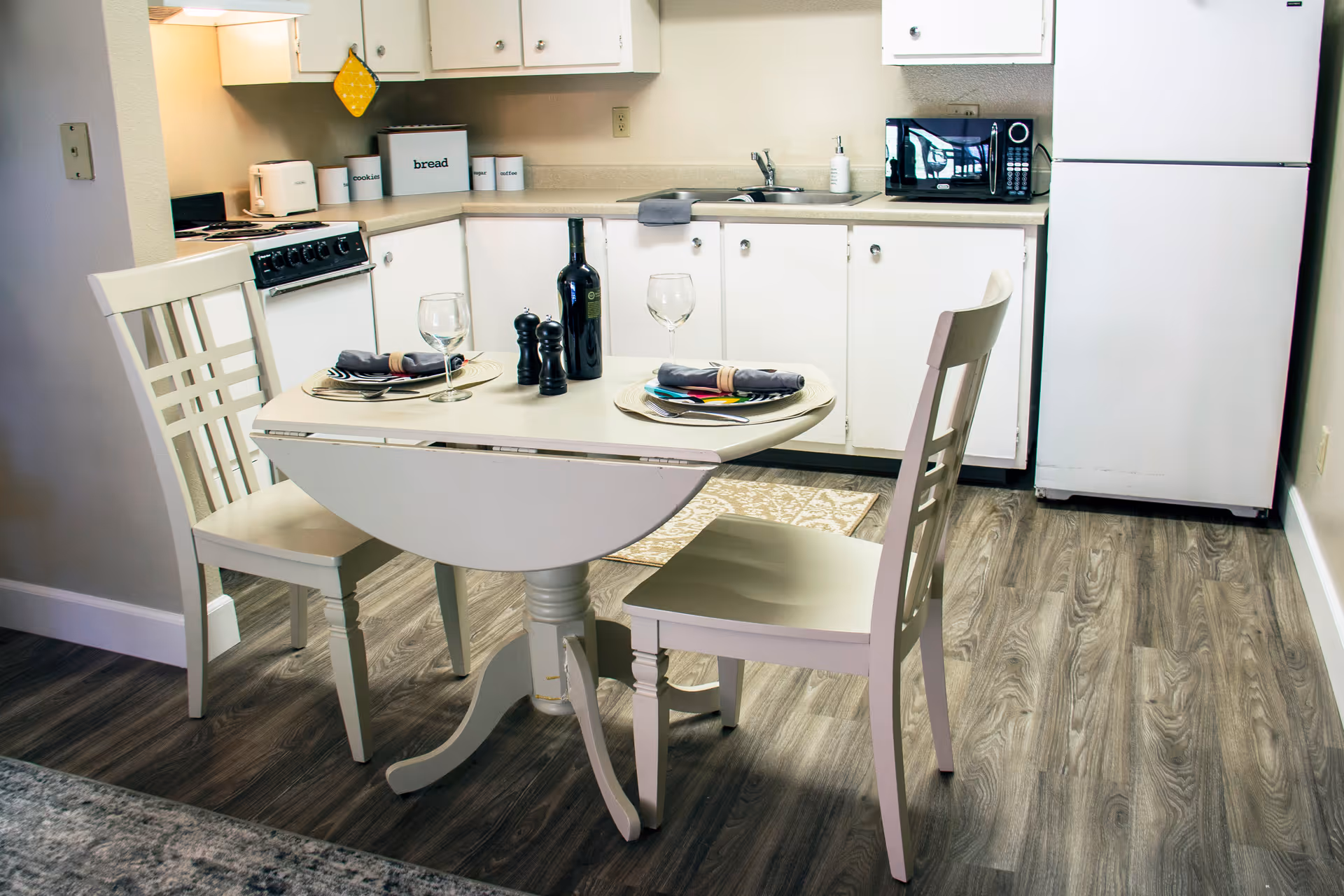 A small kitchen area with white cabinets, a white refrigerator, a microwave, and a stove. In front of the kitchen is a round white dining table set for two with wine glasses, plates, napkins, and a bottle of wine. Two white chairs are placed at the table on a wood-patterned floor.