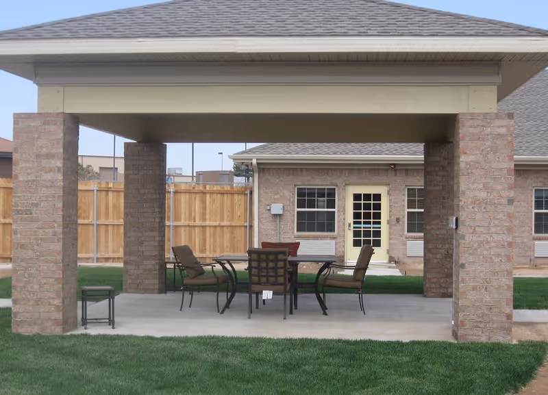 Covered outdoor patio supported by brick columns with a table and chairs on a concrete pad in front of a brick building and fenced yard.