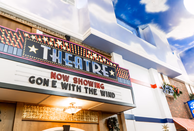Interior view of a theater entrance with a marquee sign that reads 'THEATRE' and 'NOW SHOWING GONE WITH THE WIND'. The ceiling is painted with a blue sky and white clouds, and there are decorative elements including flowers and a wreath near the entrance.