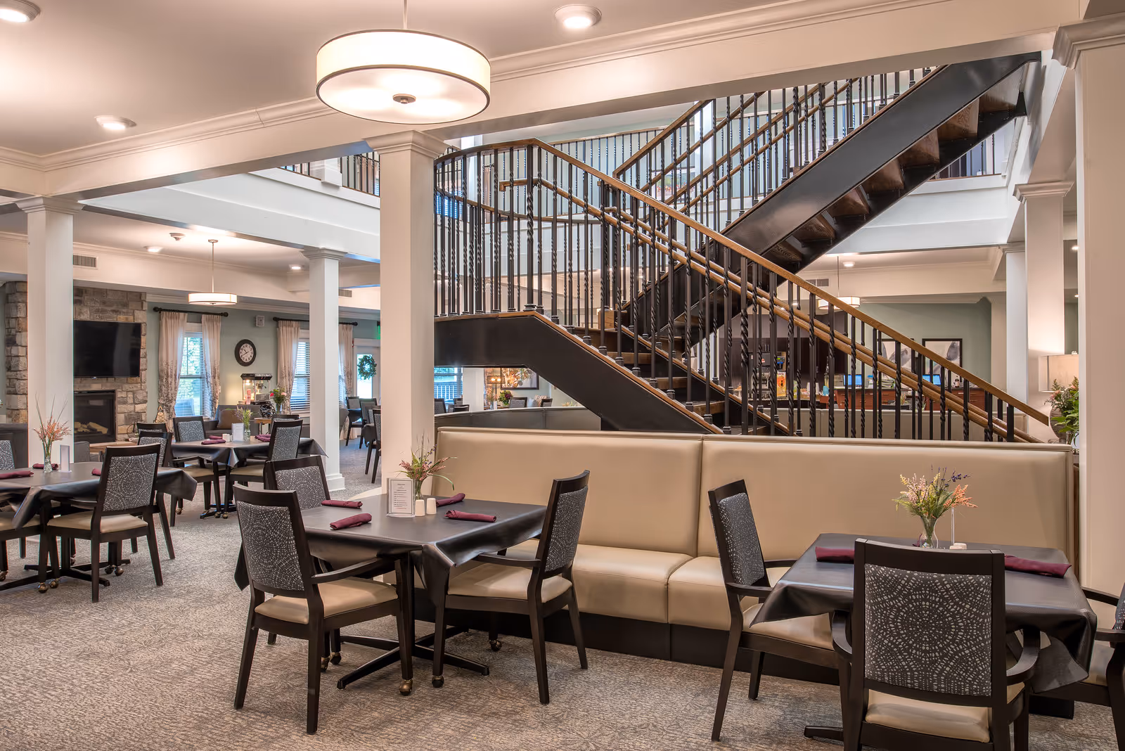 Spacious senior living dining area with tables, chairs, banquette seating and a central staircase leading to an upper level.