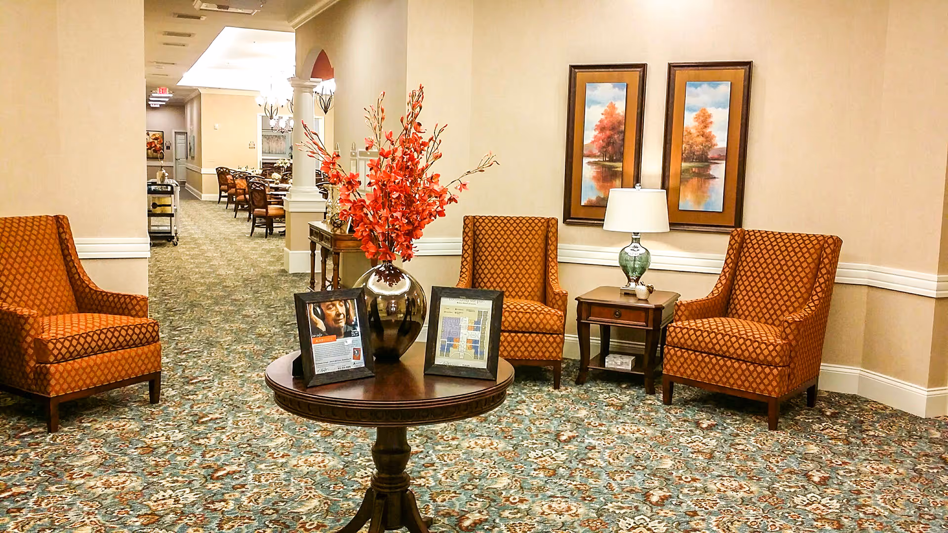 A cozy seating area in a senior living facility with three orange patterned armchairs arranged around a small wooden table with a lamp. In the foreground, a round wooden table holds a large vase with red flowers and two framed pictures. The background shows a hallway leading to a dining area with tables and chairs.