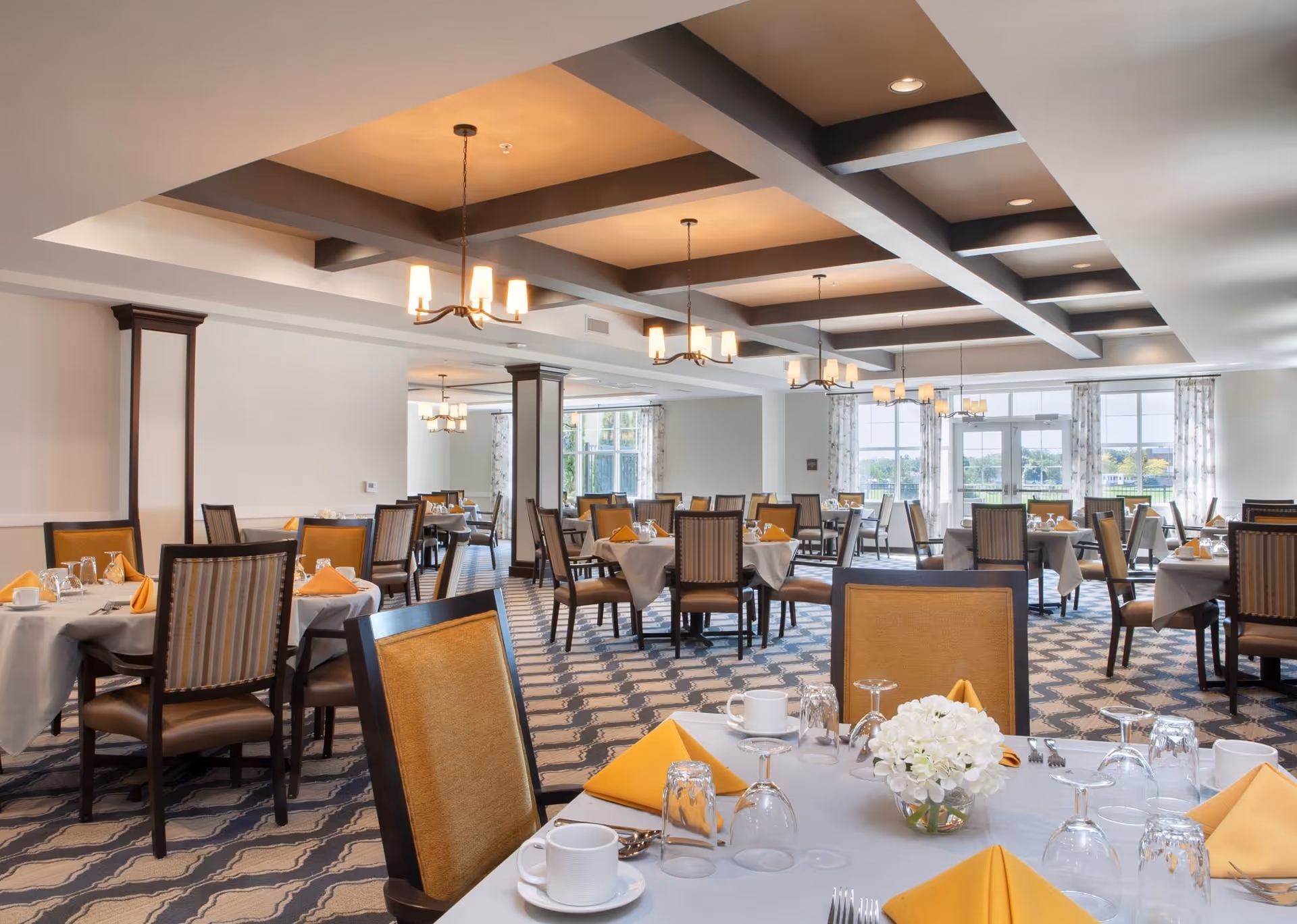 A spacious and well-lit dining room with multiple tables set with white tablecloths, yellow folded napkins, glassware, and white cups. The room features patterned carpet flooring, large windows with curtains allowing natural light, and ceiling light fixtures with multiple bulbs.