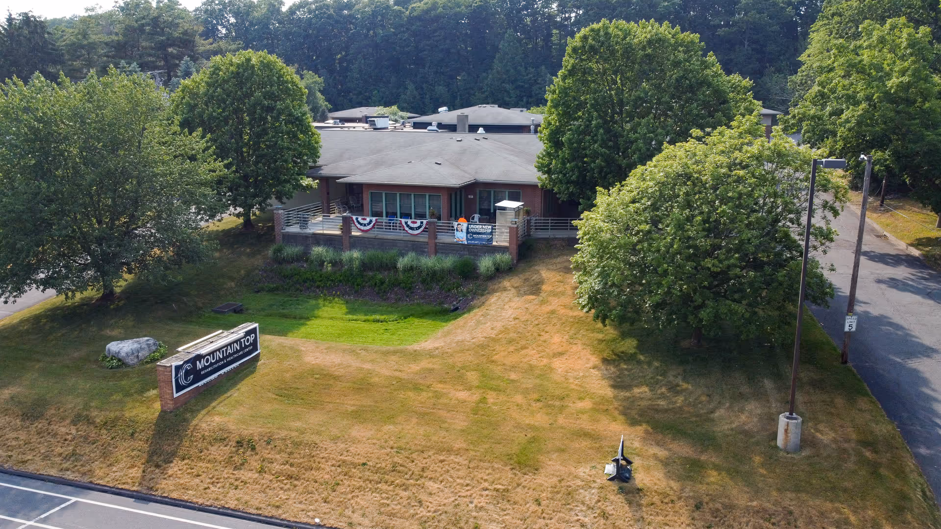 Aerial view of Mountain Top Rehabilitation and Healthcare Center building surrounded by trees and grass. The building has a low roof and a front porch decorated with patriotic bunting. There is a sign on the lawn near the road displaying the facility's name. A paved driveway and parking area are visible to the right.