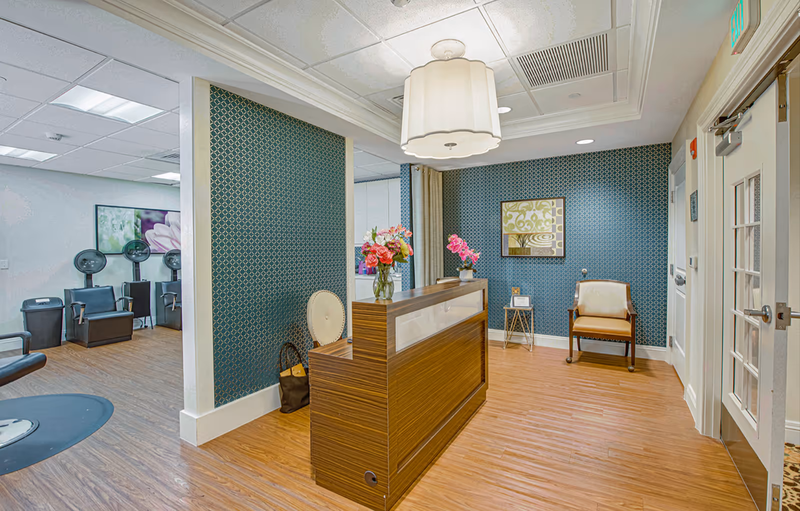 Reception area with a wooden front desk, patterned teal walls, seating, flowers on the desk, and salon chairs visible in an adjacent room.