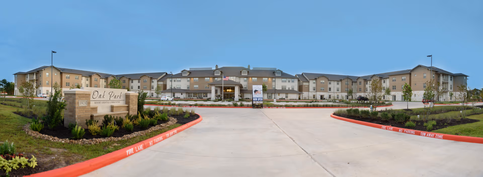 Wide exterior view of Oak Park Retirement Resort, a large multi-story building with beige and light brown walls, surrounded by landscaped greenery and a spacious driveway with red curbs marked as fire lanes.