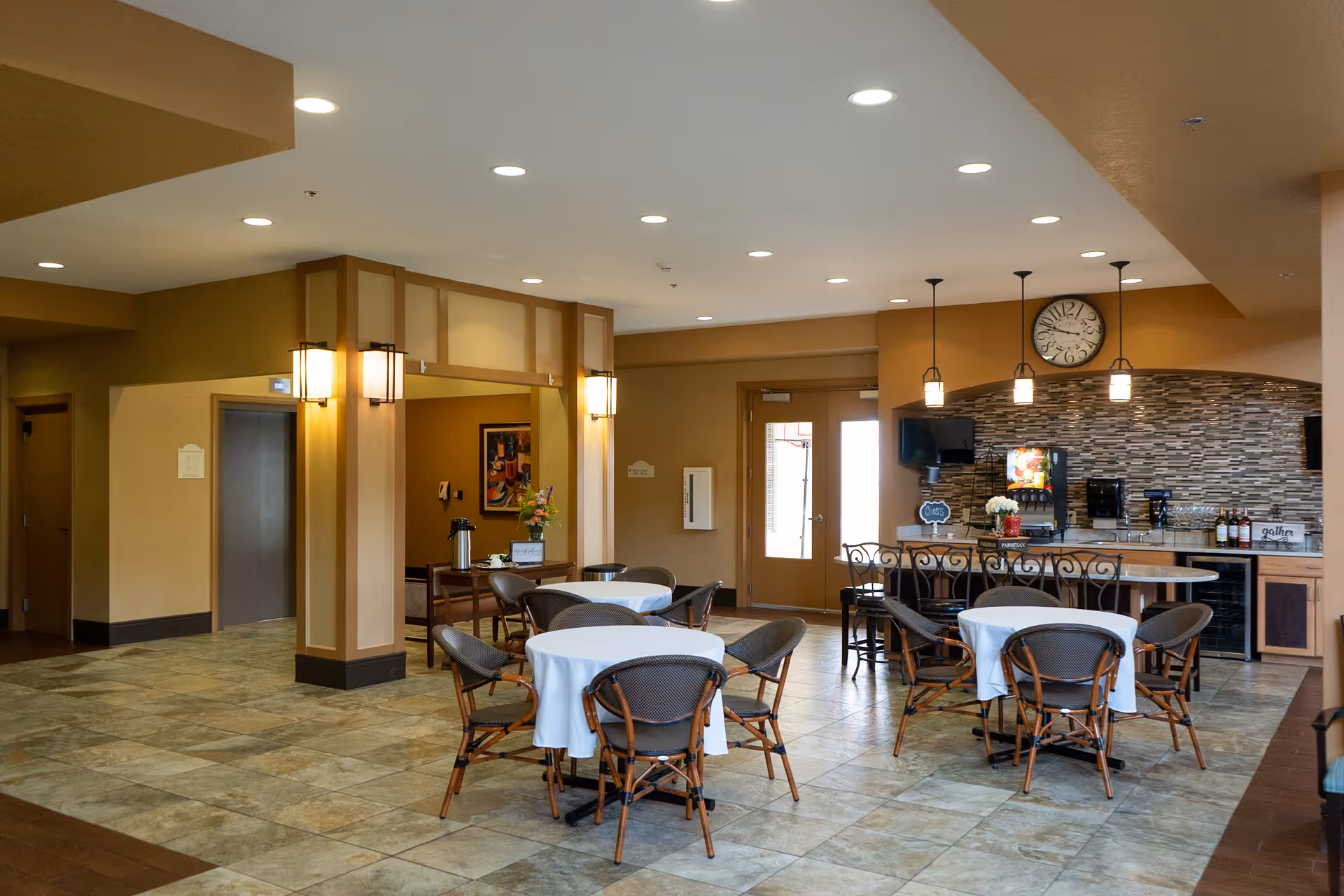 Spacious dining area with round tables covered in white tablecloths, wicker chairs, and a counter with pendant lights and a clock on the wall.