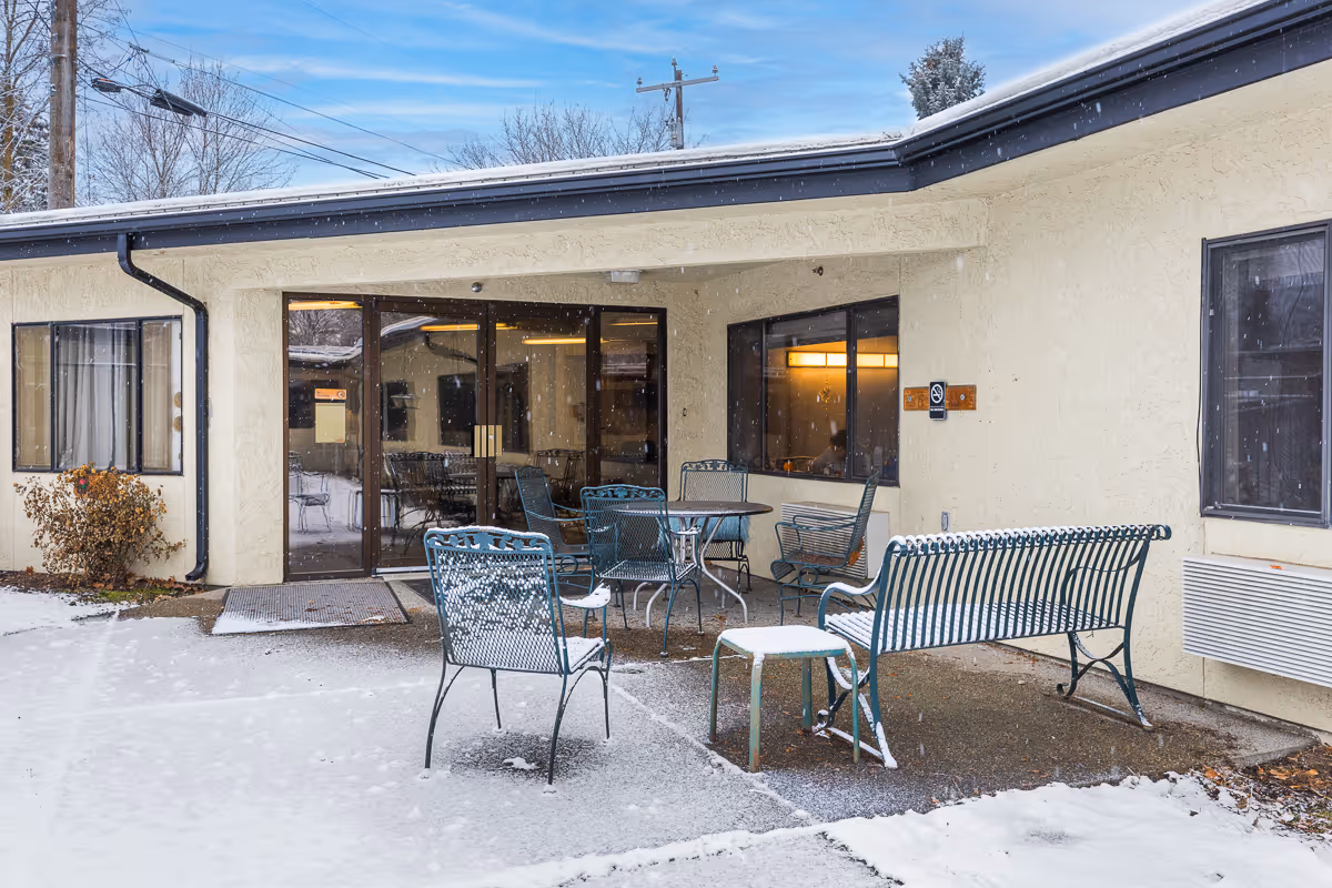 Outdoor patio area of a senior living facility with metal chairs, benches, and tables lightly covered in snow. The building has beige stucco walls and large windows, with a glass door leading inside. Snow is falling and covering parts of the ground and furniture.