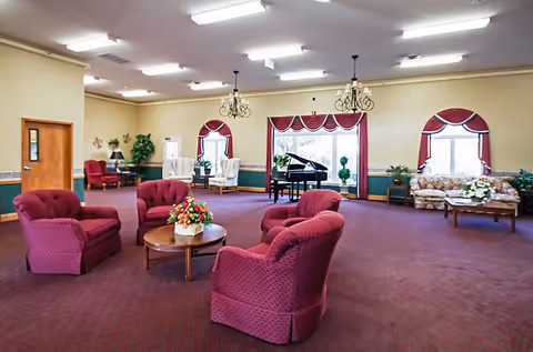 Spacious senior living common room with red upholstered chairs around a coffee table, chandeliers, a grand piano, and windows with red valances.