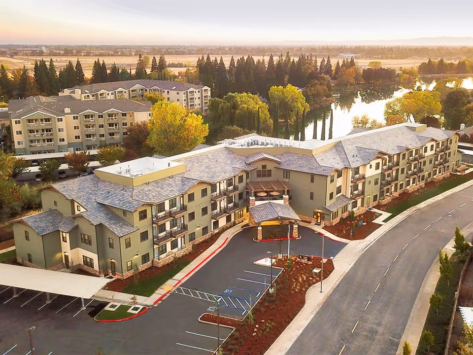 Aerial view of Westmont Of Chico - The Inn, a multi-story senior living facility surrounded by trees and a pond, with a parking lot and a curved road in front of the building during sunset.