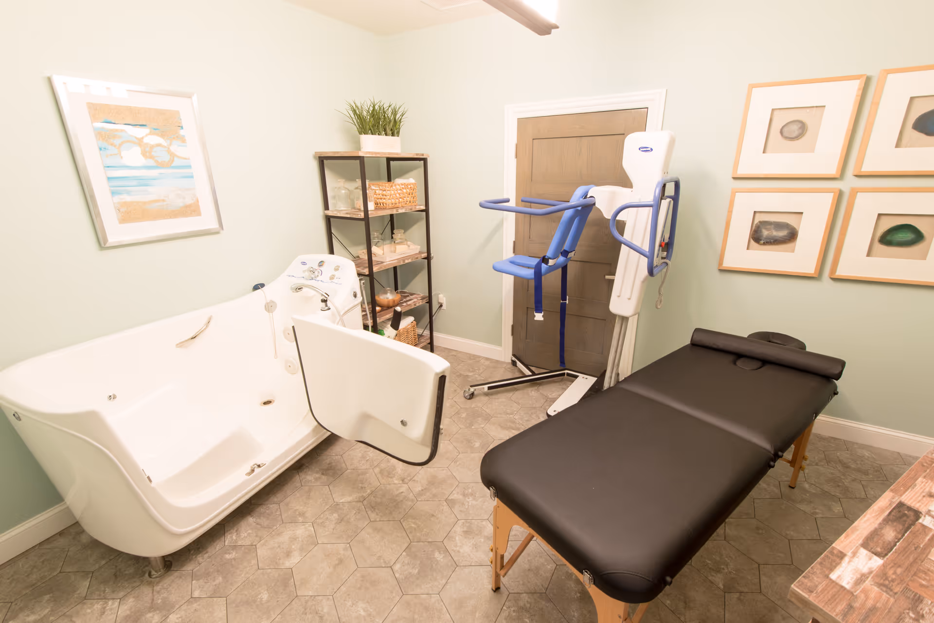 A bathroom with a walk-in bathtub featuring a side door, a black massage table, a patient lift device, a shelving unit with towels and baskets, and framed artwork on the walls.