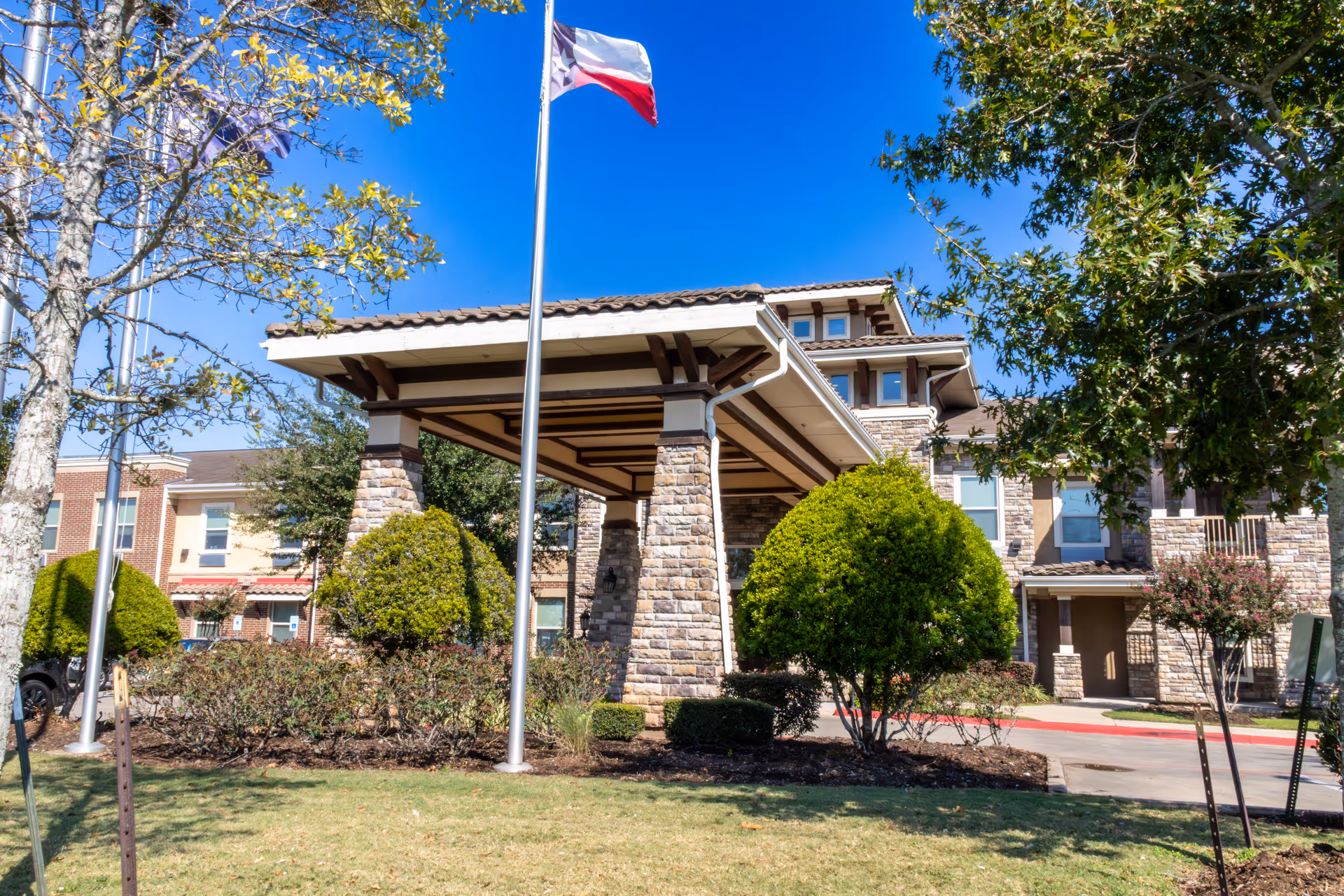 Exterior view of a senior living facility with a covered entrance supported by stone pillars, manicured bushes, trees, and a flagpole with a flag flying against a clear blue sky.