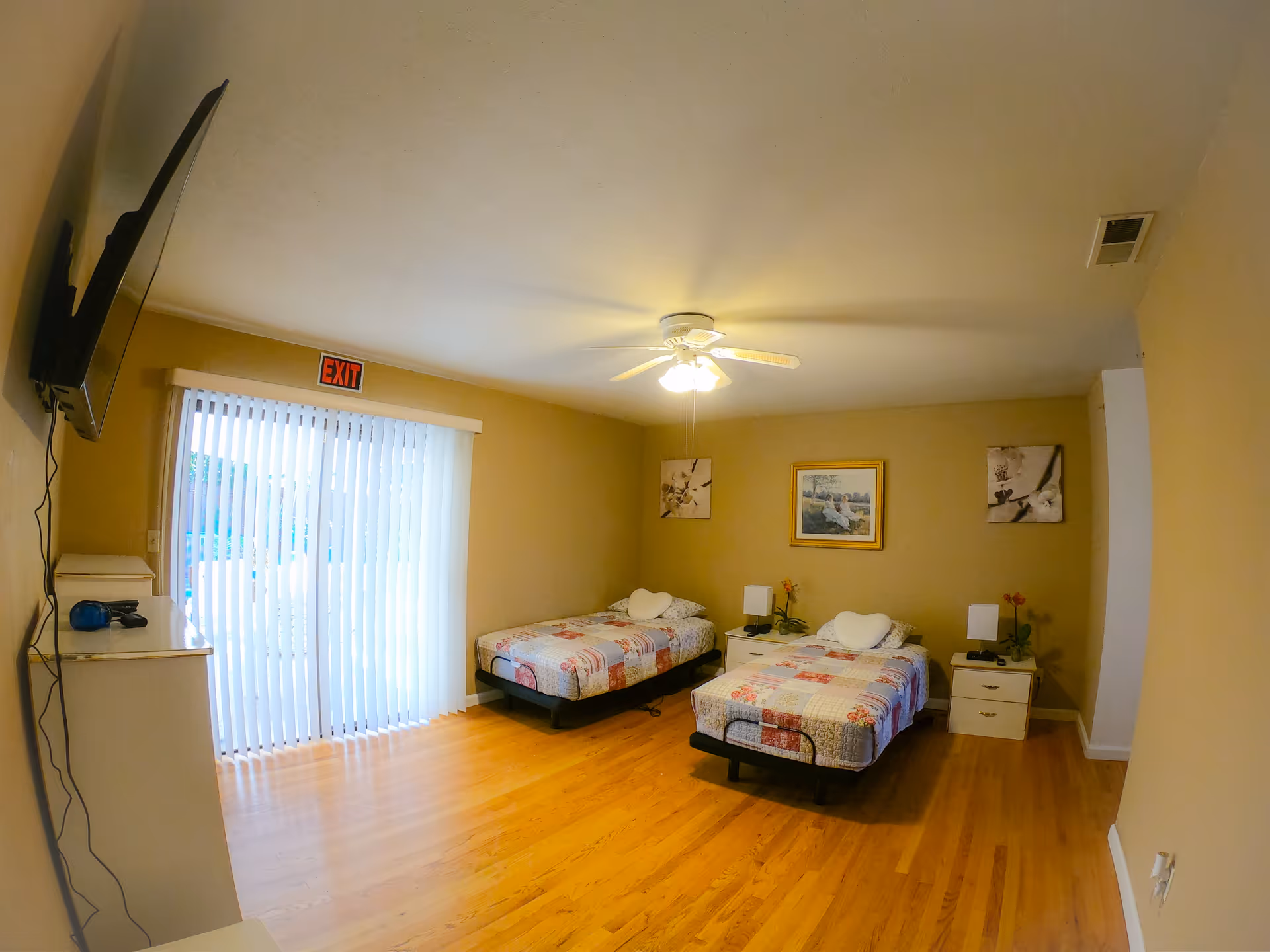 Small senior living bedroom with two single beds, nightstands, ceiling fan, wall-mounted TV, and a sliding glass door with vertical blinds.