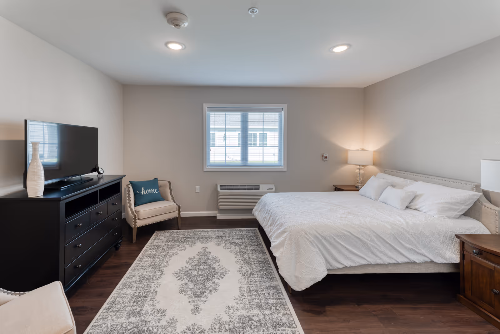 A bright and clean bedroom in a senior living facility featuring a large bed with white bedding, two wooden nightstands with lamps, a patterned area rug, a black dresser with a flat-screen TV and decorative vase, a beige armchair with a blue pillow that says 'home', and a window with blinds letting in natural light.