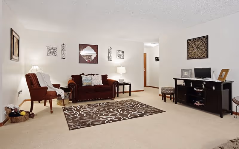 A cozy living room with a maroon upholstered sofa and matching armchair, a patterned area rug on a beige carpet, decorative wall art, a side table with a lamp, and a dark wooden console table with framed pictures and a small TV.