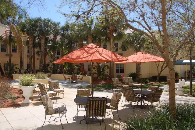 Sunlit courtyard with round tables, wrought-iron chairs and red umbrellas surrounded by palm trees and the facility building.